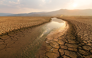 A river which appears to be gradually drying up with cracked, dry earth next to it. The sun setting over mountains is in the background.